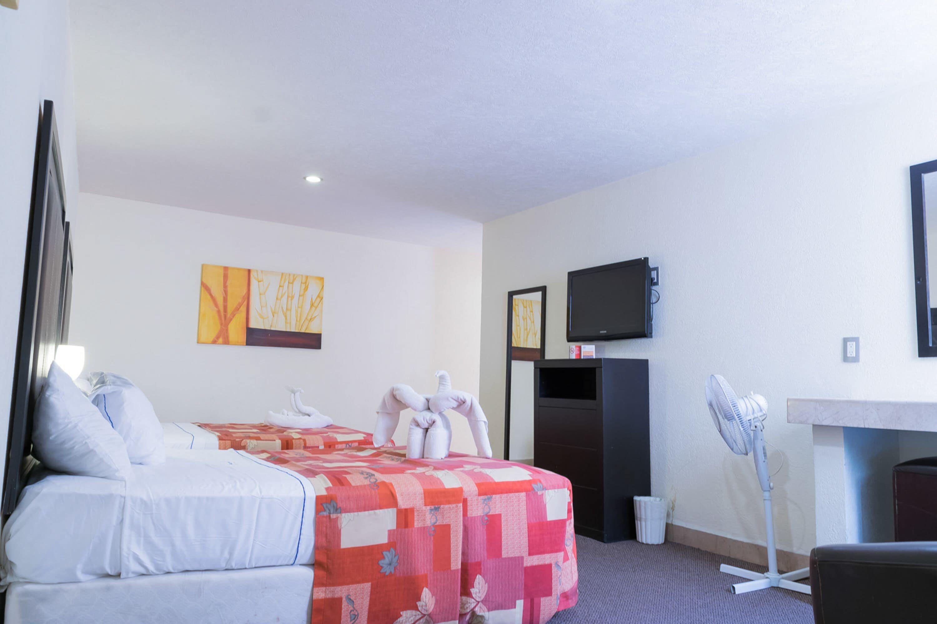 A hotel room with a bed, a desk, a chair, a standing fan, a TV, and artwork on the wall.