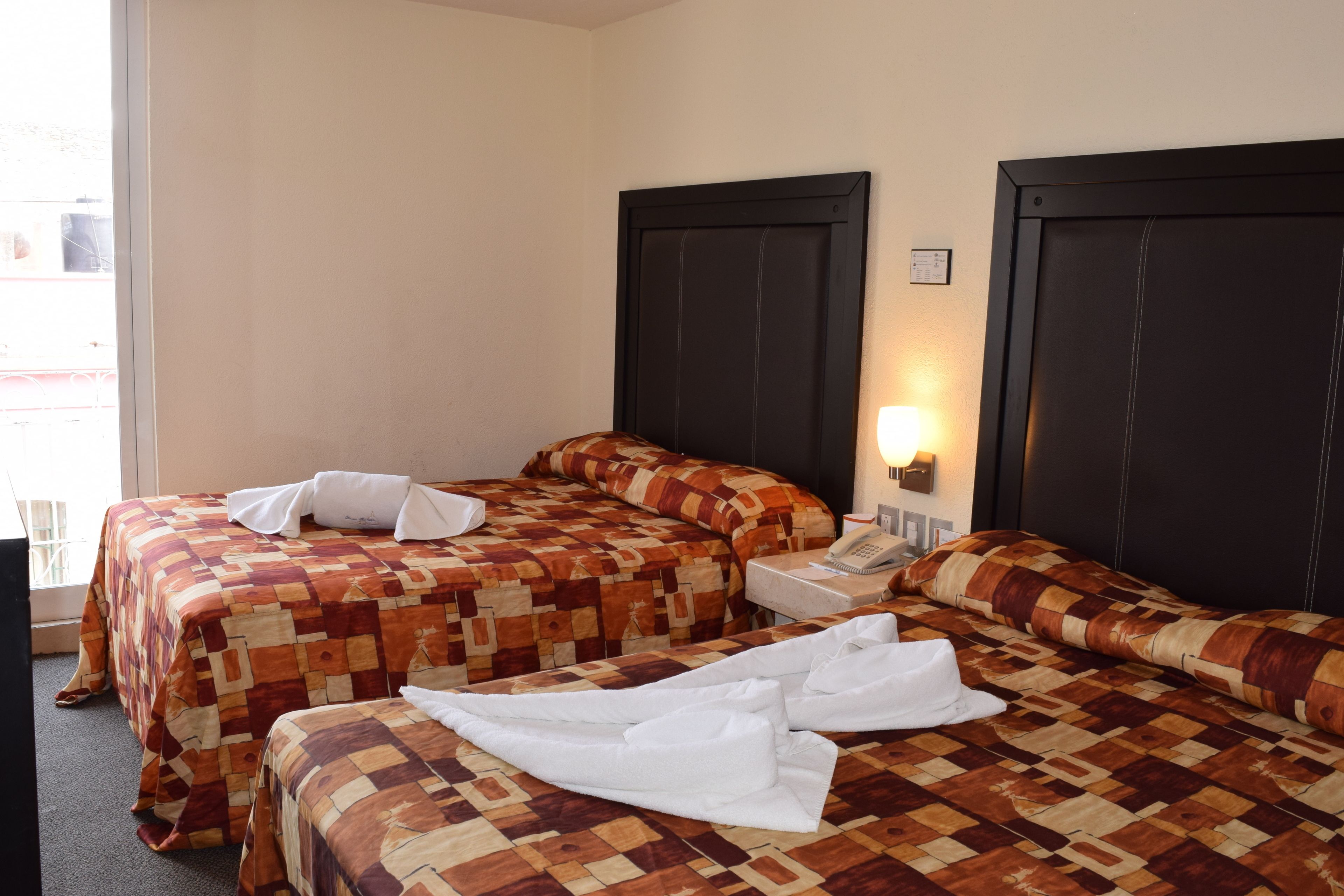 A hotel room with two beds, each with a quilted bedspread and a white pillow. There is a bedside table with a lamp and a telephone.