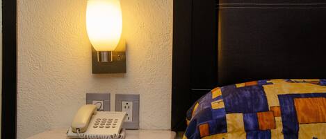 A hotel room with a marble bedside table, a telephone, a wall-mounted light, and a bed with a patterned blanket.