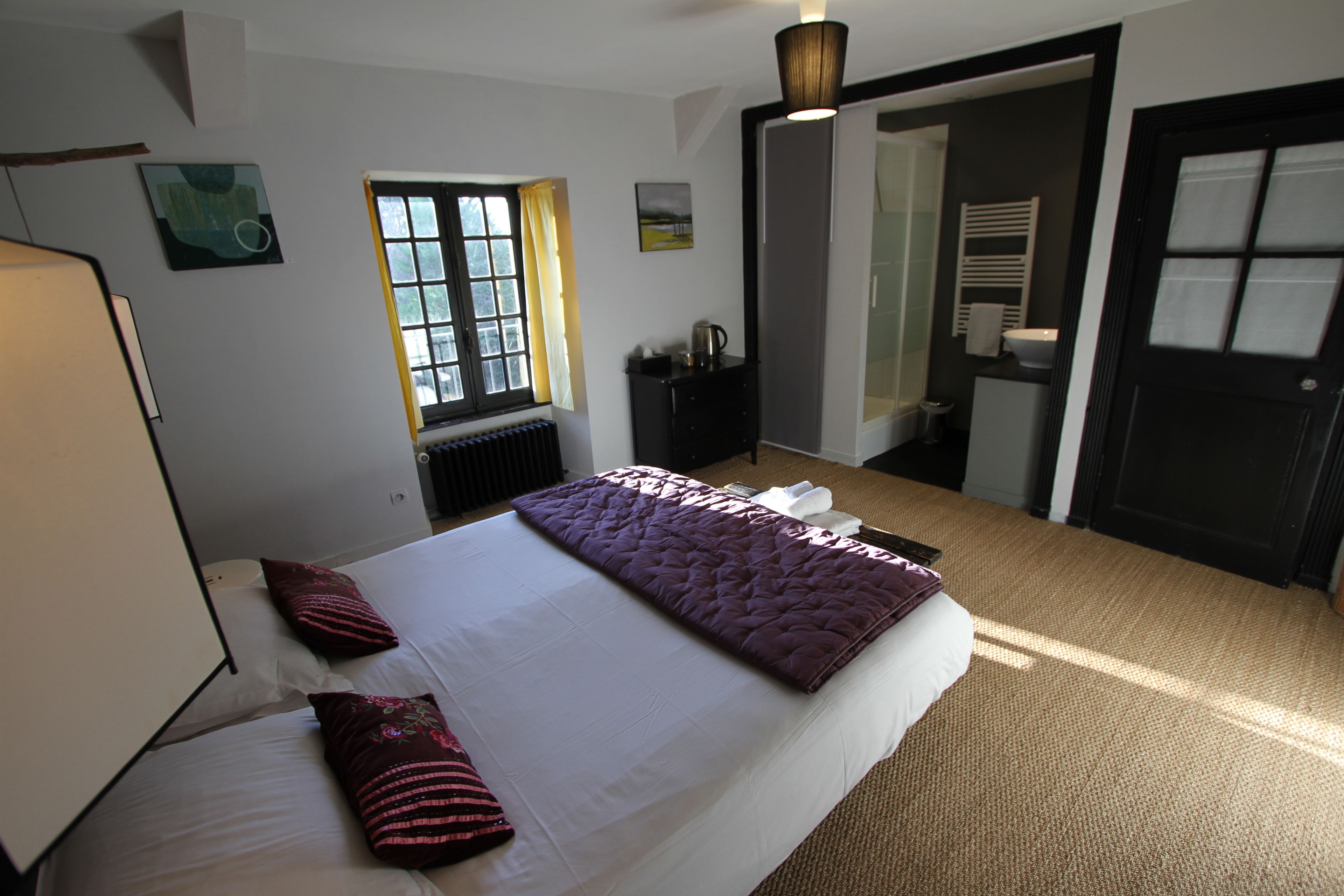 Habitación de hotel con una cama grande, una ventana con cortinas, un radiador, un armario pequeño y una puerta que da a otra habitación.