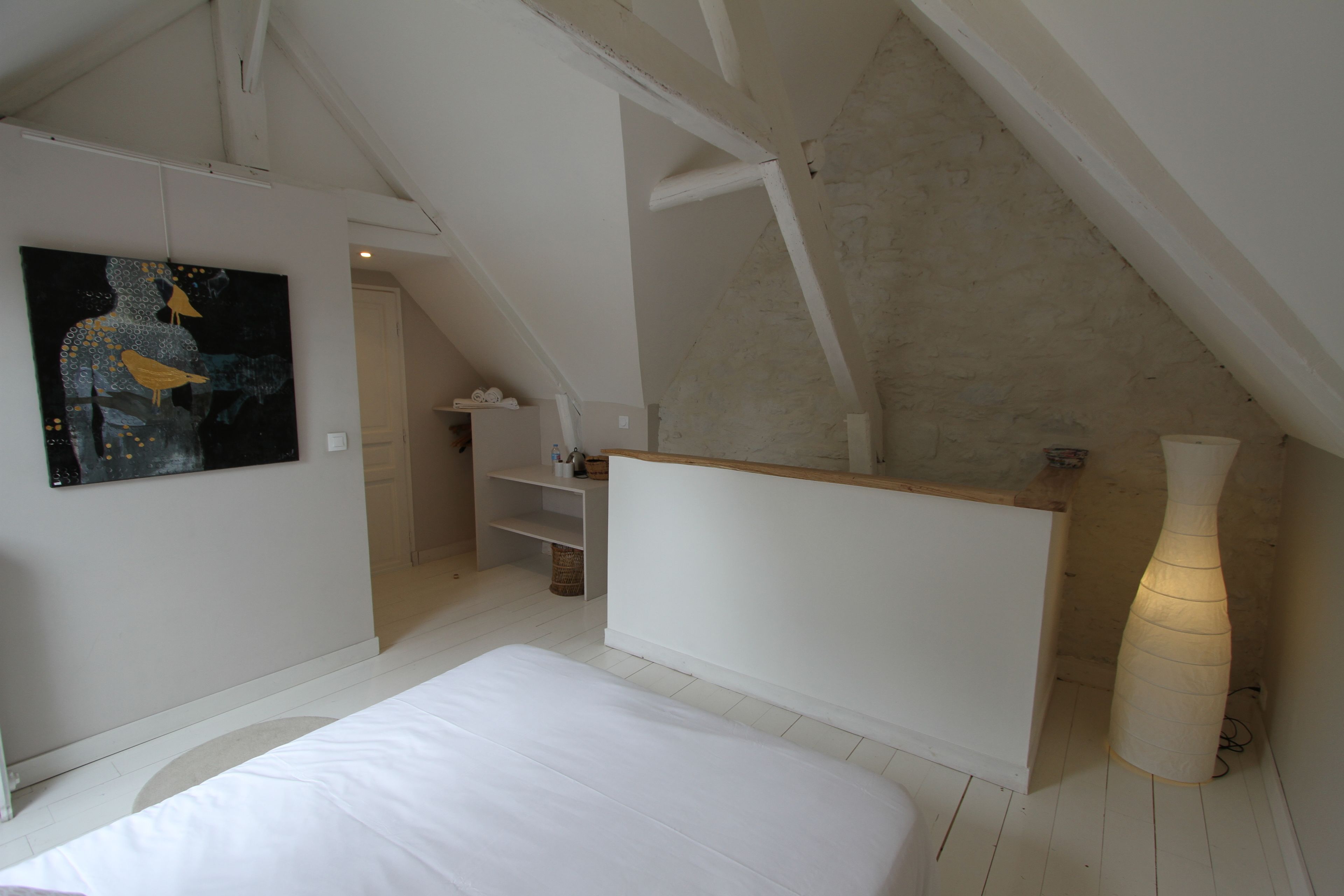 Une chambre avec un plafond en pente, un lit, un tableau, une table de chevet et un grand vase.