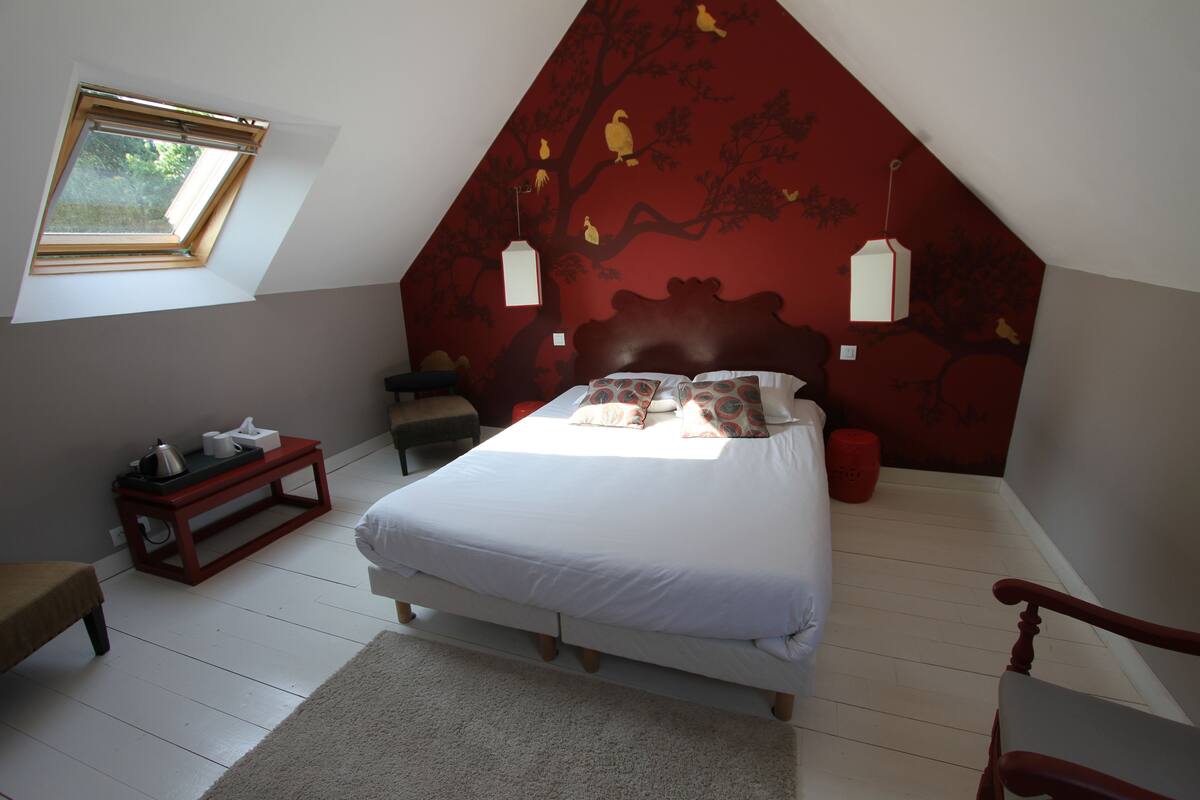 A bedroom with a sloped ceiling, a bed with white bedding, a red wall with a tree mural, a small table with a kettle, and a window with a view of greenery.