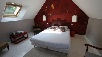 Un dormitorio con techo inclinado, una cama con ropa de cama blanca, una pared roja decorada con un mural de un árbol, una mesita con un hervidor y una ventana con vistas a follaje.