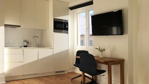 Une cuisine compacte avec des meubles blancs, un four à micro-ondes et un évier. On y trouve une table à manger en bois, une chaise et un téléviseur à écran plat fixé au mur.