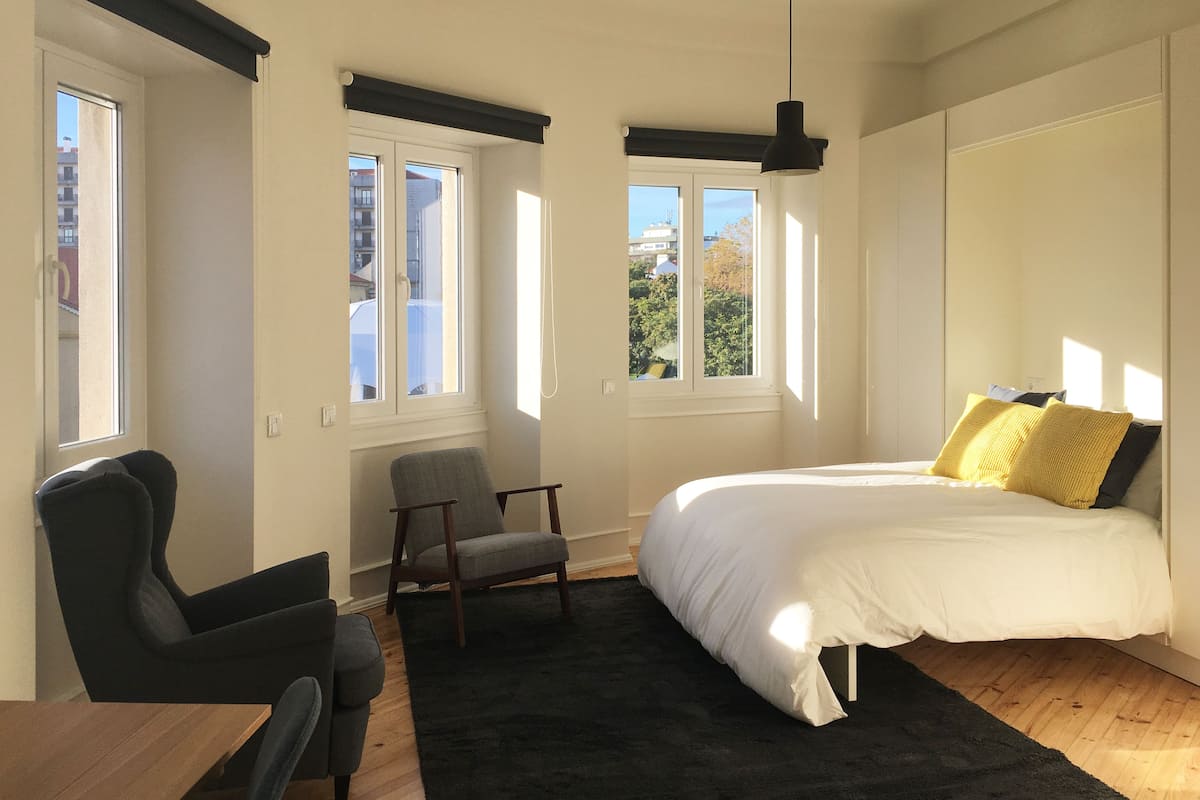 A bedroom with a bed, a chair, a desk, and two windows.