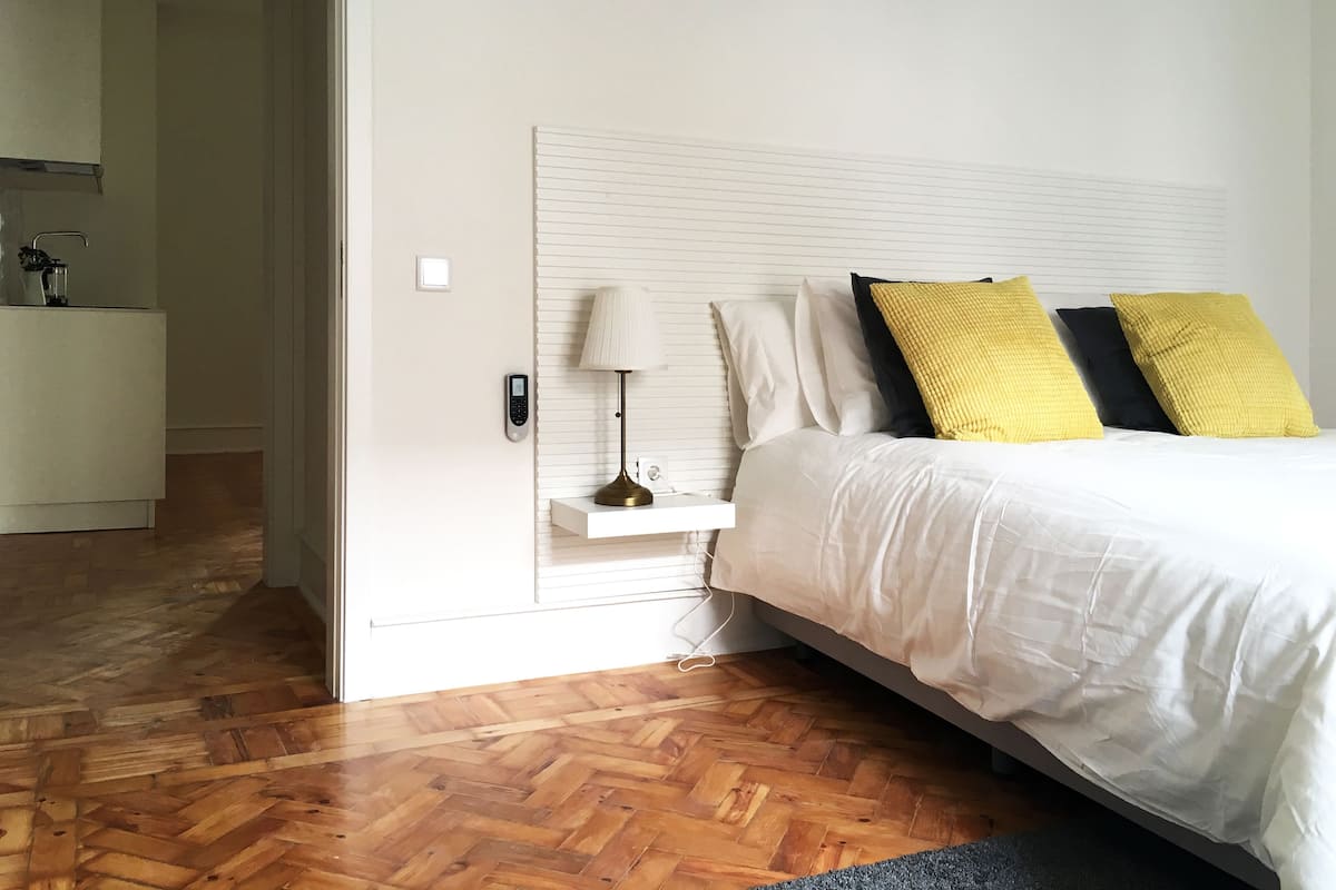 A bedroom with a white headboard, a bedside table with a lamp, and a bed with white bedding and two pillows.