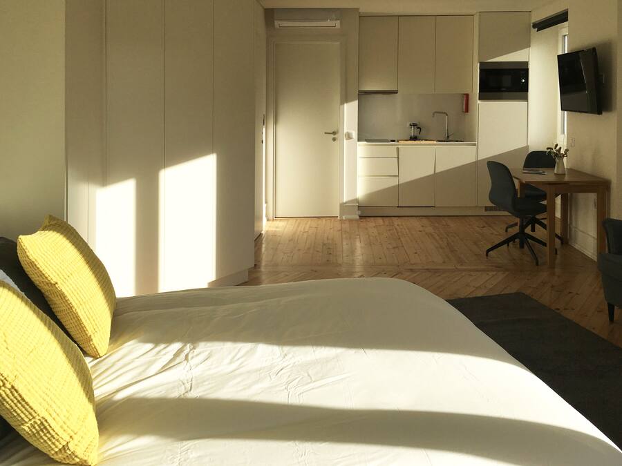 A bedroom with a bed, a desk, and a kitchenette in the background.