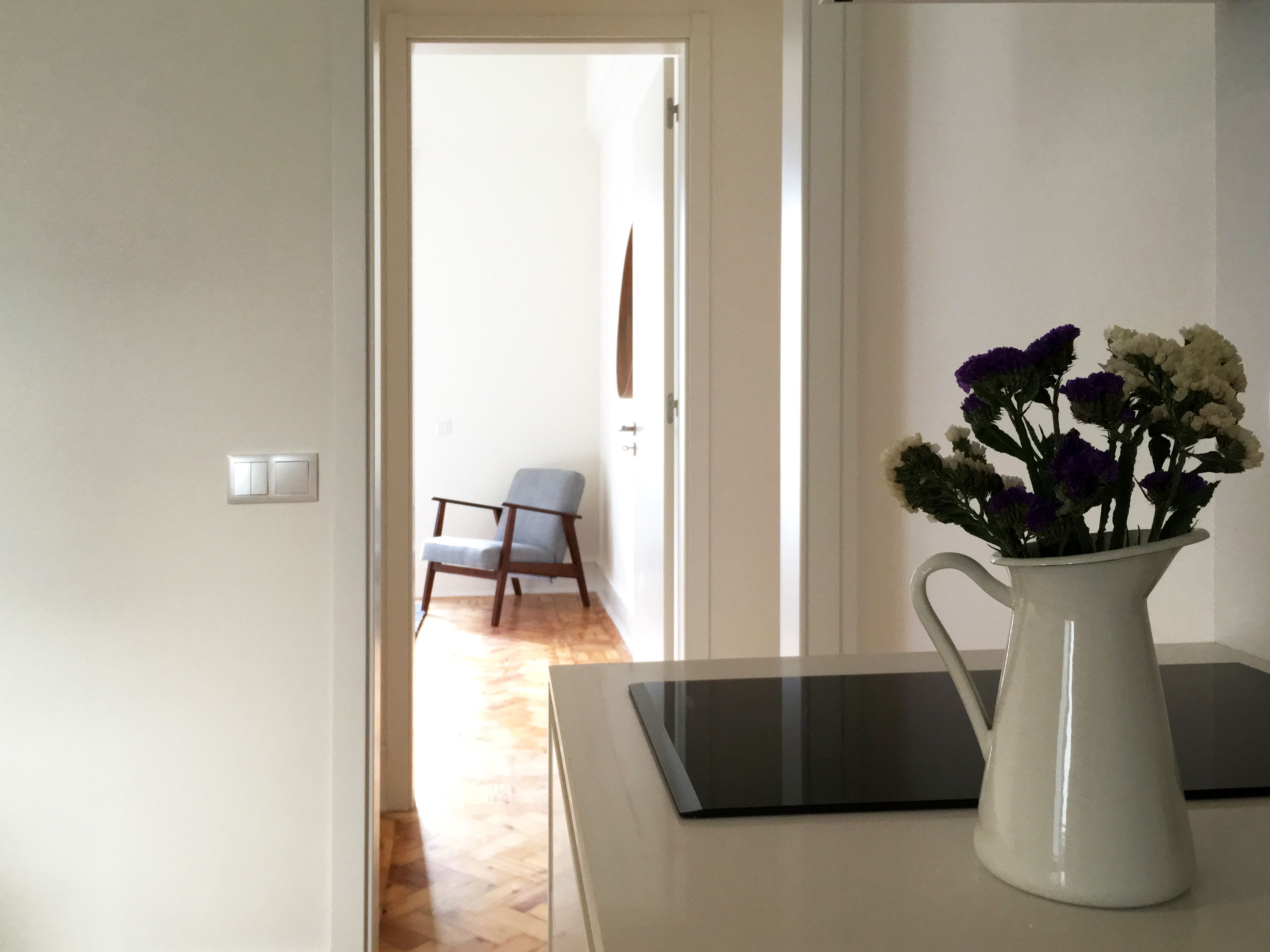 A room with a white wall, a wooden floor, a white vase with flowers, and a black tablet on a white table.