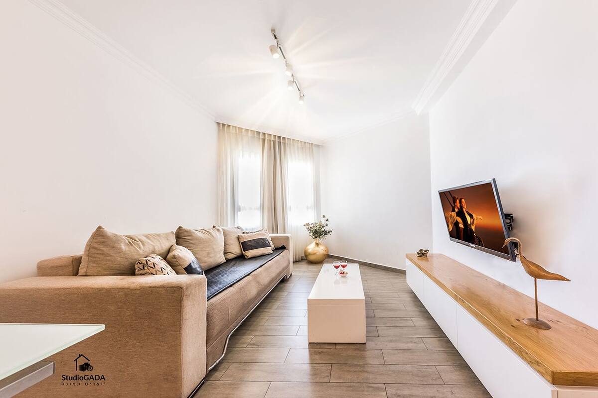 A modern living room with a sofa, a coffee table, a TV mounted on the wall, and a vase with flowers.