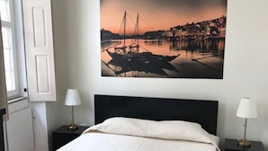 A neatly made bed with a white comforter and two rolled towels, a wall-mounted painting of a sunset harbor, and two bedside tables with lamps. - Linha22 GuestHouse (Porto)