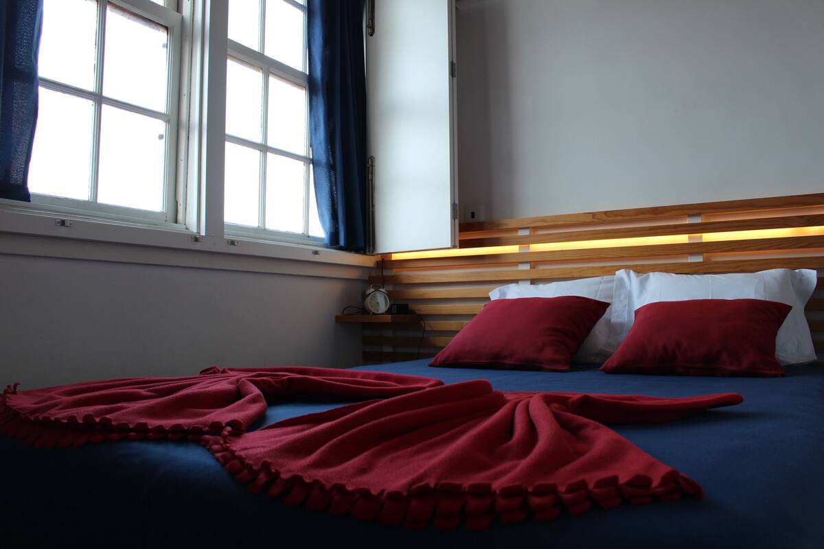 A bedroom with a bed, red pillows, a wooden headboard, a bedside table with a clock, and a window with curtains.