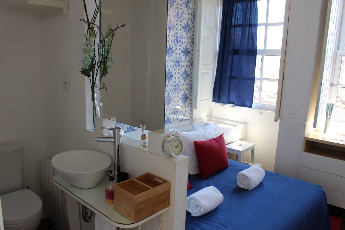 A bathroom with a white sink, a mirror, a clock, a wooden tray, and a bed with blue bedding and red pillows.