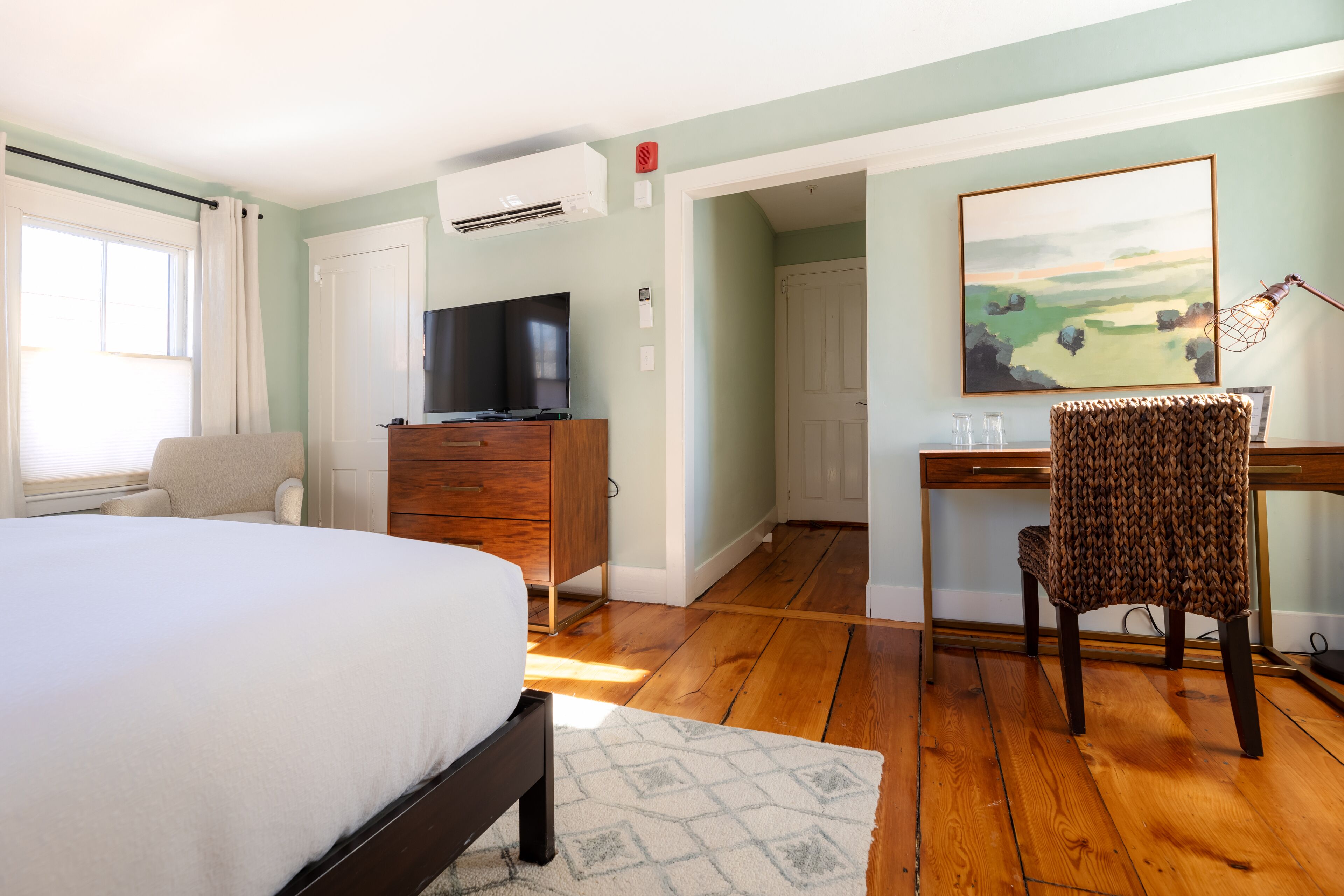 A bedroom with a bed, dresser, TV, desk, chair, and a painting on the wall.