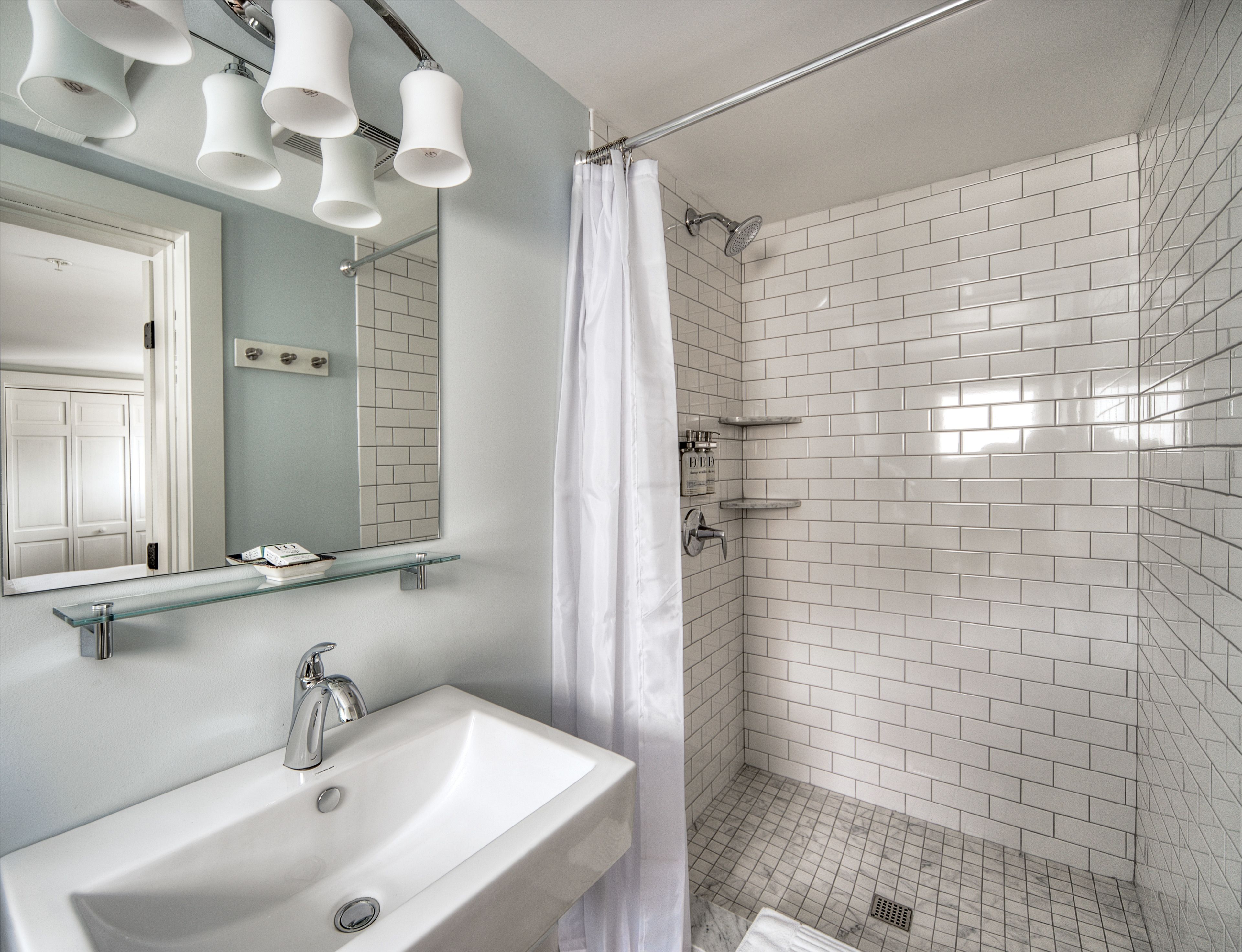 Casa de banho com lavatório branco, espelho e chuveiro com azulejos brancos.