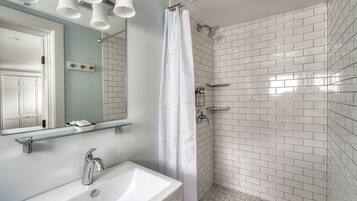 Casa de banho com lavatório branco, espelho e chuveiro com azulejos brancos.