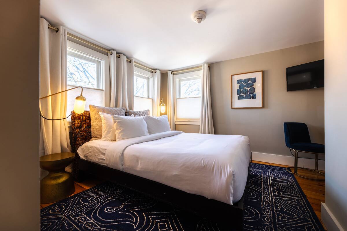 A bedroom with a bed, a side table, a lamp, a chair, a TV, and a window with curtains.