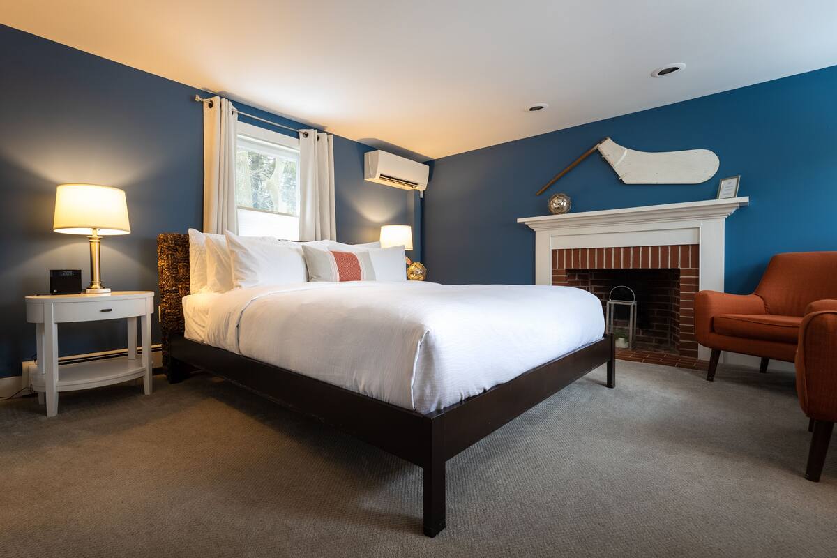 A bedroom with a bed, two bedside tables, a chair, a fireplace, and an air conditioning unit.