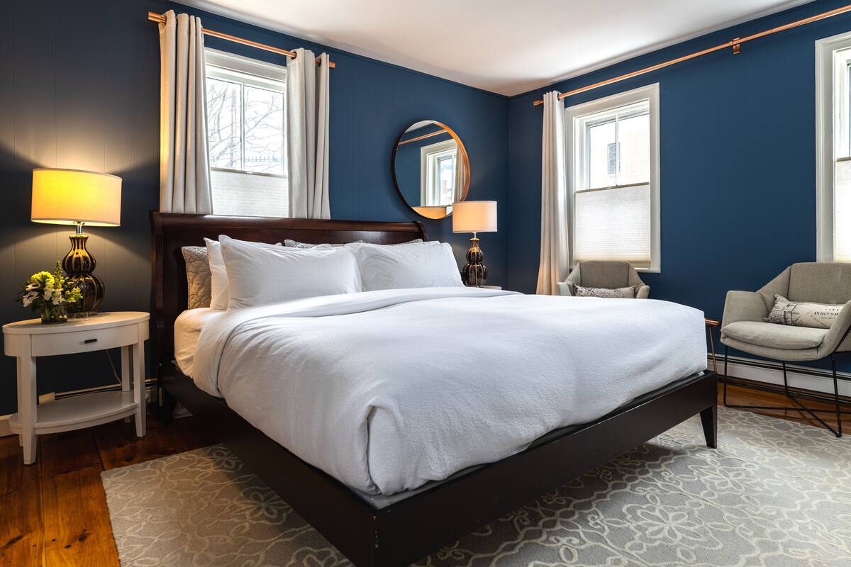 A neatly made bed in a room with dark blue walls, two bedside tables with lamps, a chair, and a large mirror on the wall.