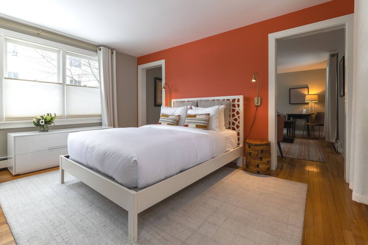 A modern bedroom with a white bed, a wooden nightstand, and a view of the hallway.