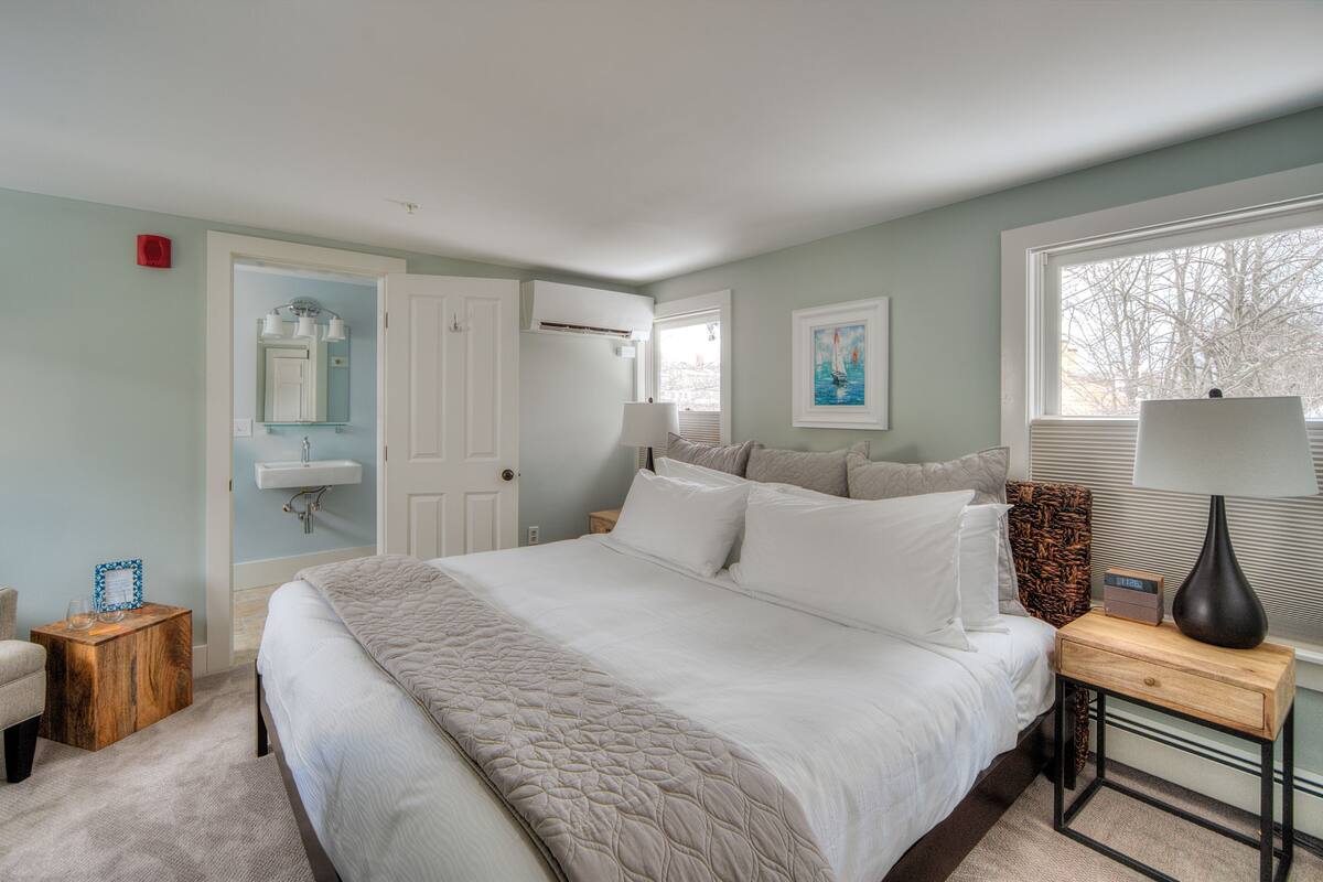 A bedroom with a large bed, bedside tables, a chair, a window with blinds, and a door leading to another room.