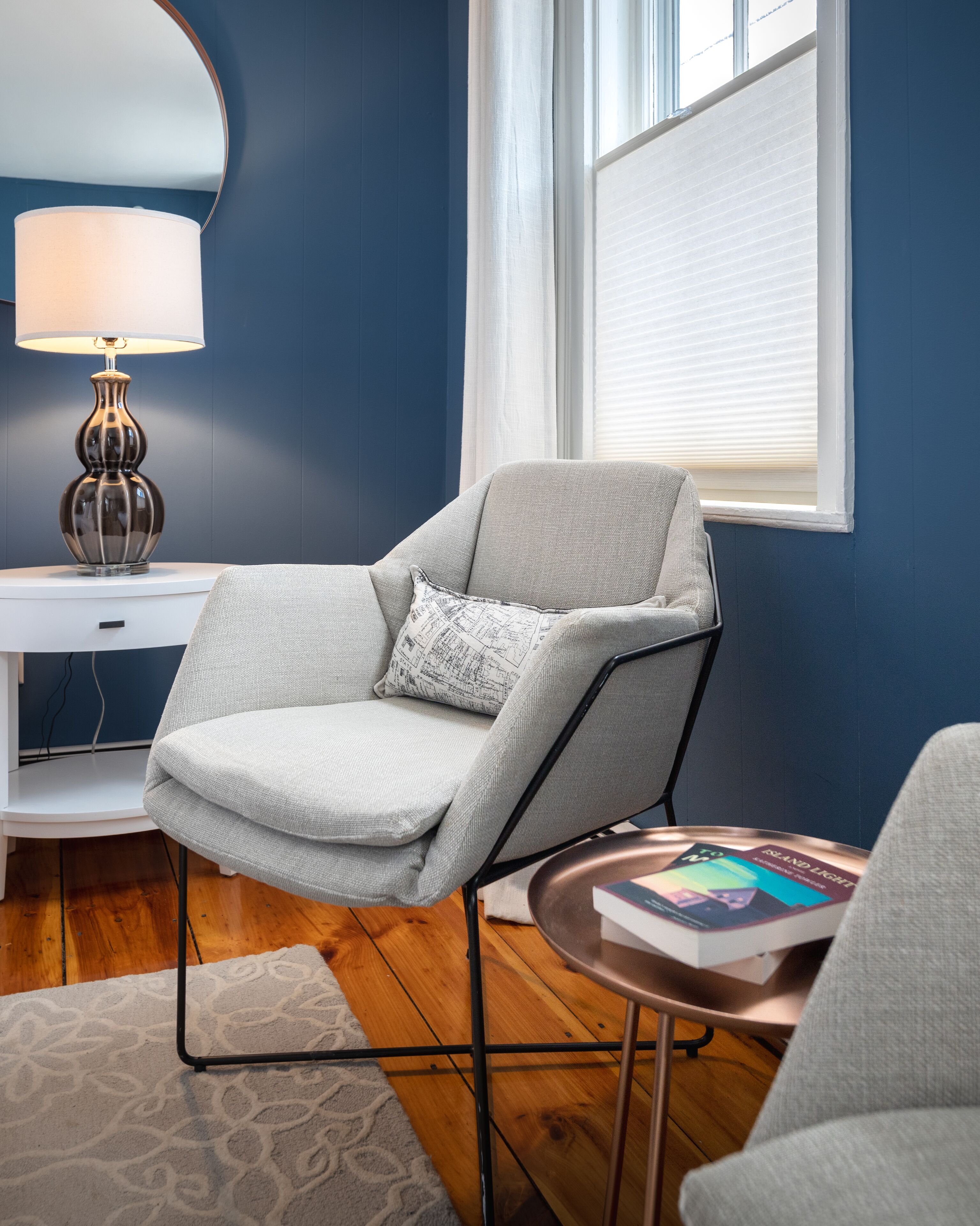 Una sala de estar moderna con un sillón gris, una mesita auxiliar con un libro, una lámpara y una ventana con persianas.