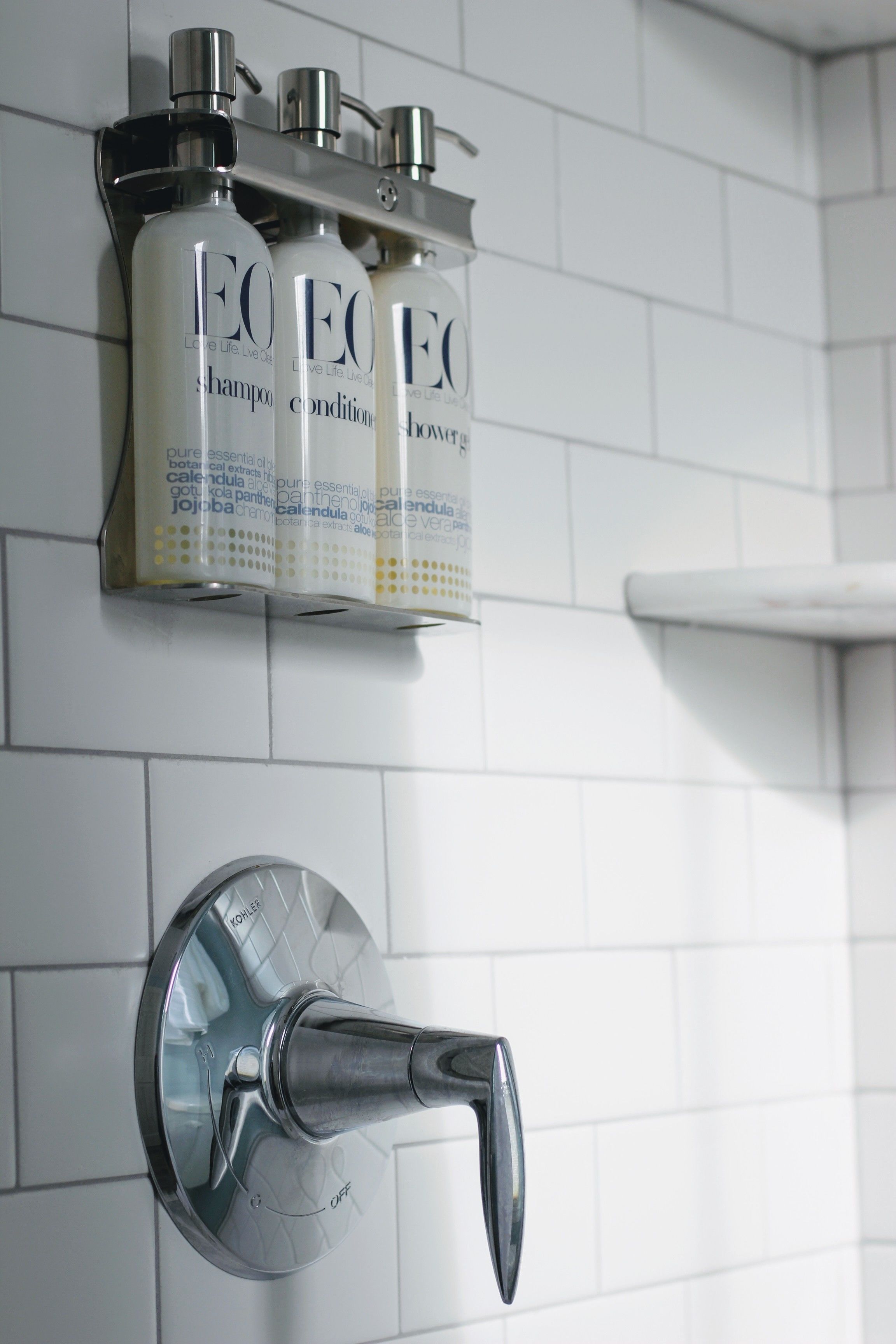 Une salle de bain équipée d’un distributeur mural pour shampoing et après-shampoing, d’un robinet Kohler et d’un mur recouvert de carreaux blancs.