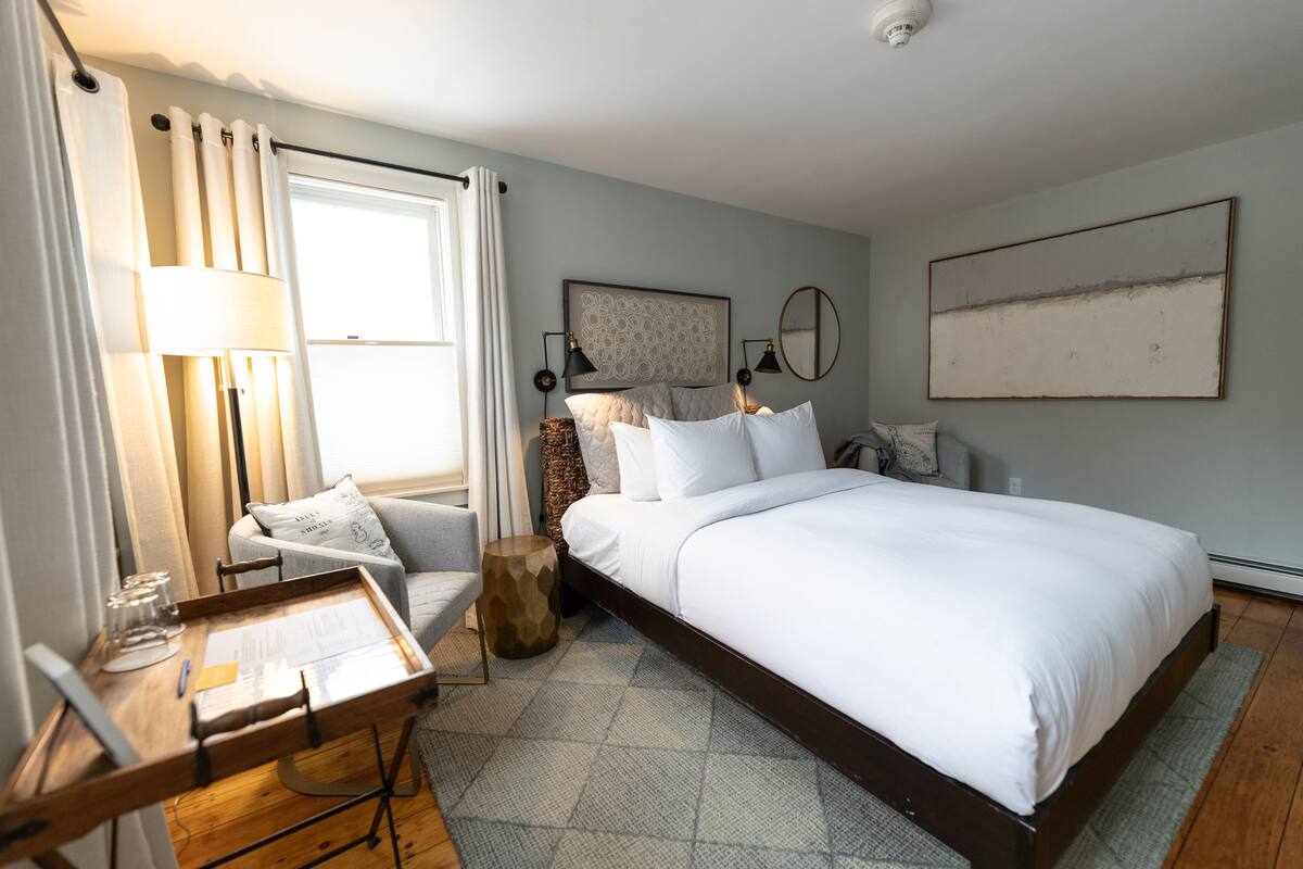 A bedroom with a large bed, a bedside table, a chair, a lamp, and a window with curtains.