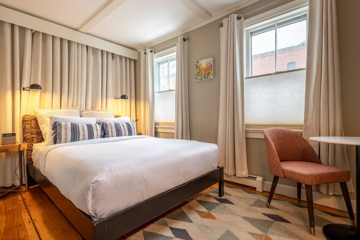 A neatly arranged bedroom with a bed, bedside lamps, a chair, and a window with curtains.