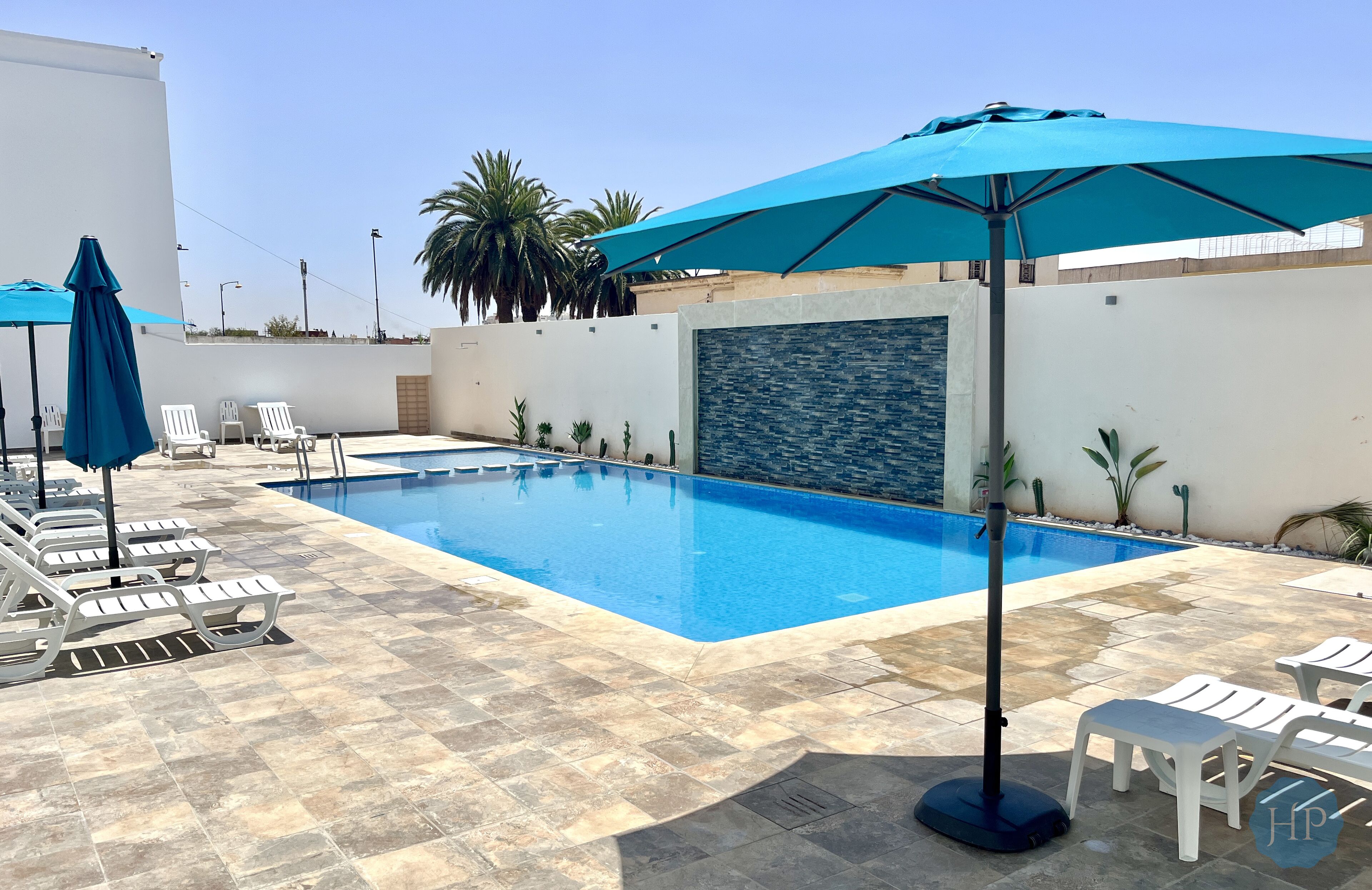 A swimming pool with lounge chairs and umbrellas, surrounded by a stone-tiled patio and palm trees.