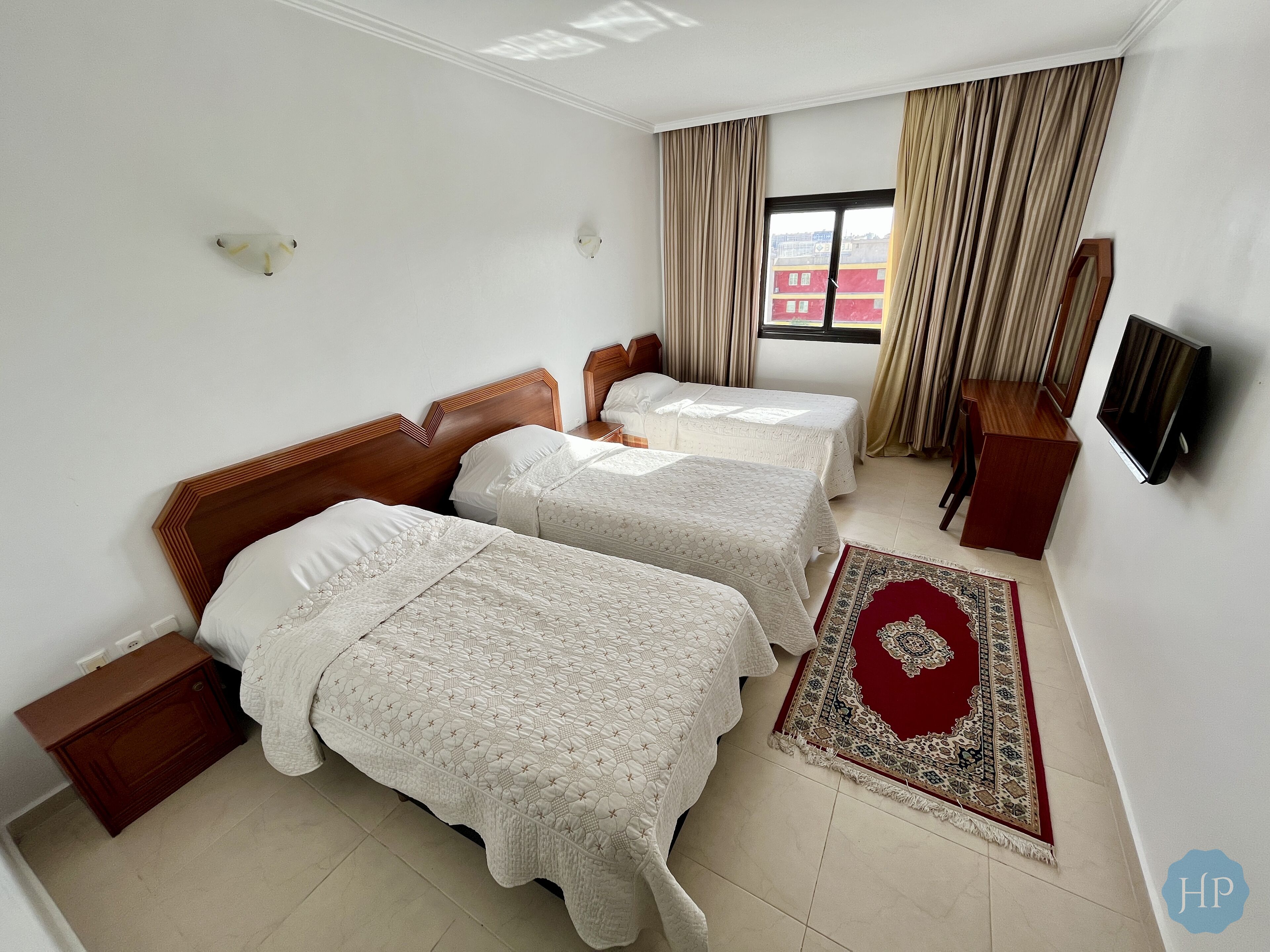 A hotel room with two beds, a TV, a wooden nightstand, a red and beige rug, and a window with curtains.
