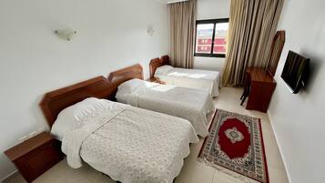 A hotel room with two beds, a TV, a wooden nightstand, a red and beige rug, and a window with curtains.