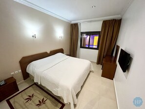 Habitación de hotel con una cama grande, una mesita de noche, un televisor montado en la pared y una ventana con cortinas.