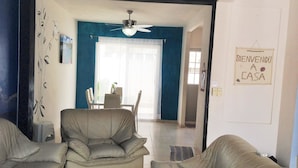 Una sala de estar con dos sofás color beige, un ventilador de techo y una zona de comedor con mesa y sillas. Se ve un letrero en la pared que dice 'BIENVENIDO A CASA'.