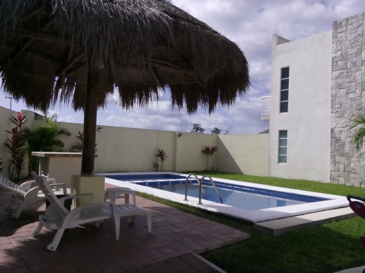 A swimming pool with a thatched roof, a white building with stone accents, and a patio area with chairs and plants.