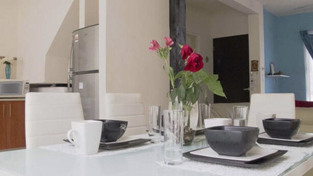 Comedor con una mesa blanca dispuesta para cuatro, incluyendo platos, tazas y un florero con flores.