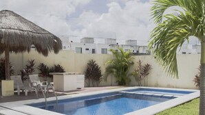 Una piscina rectangular con sombrilla de paja, rodeada de césped y palmeras.