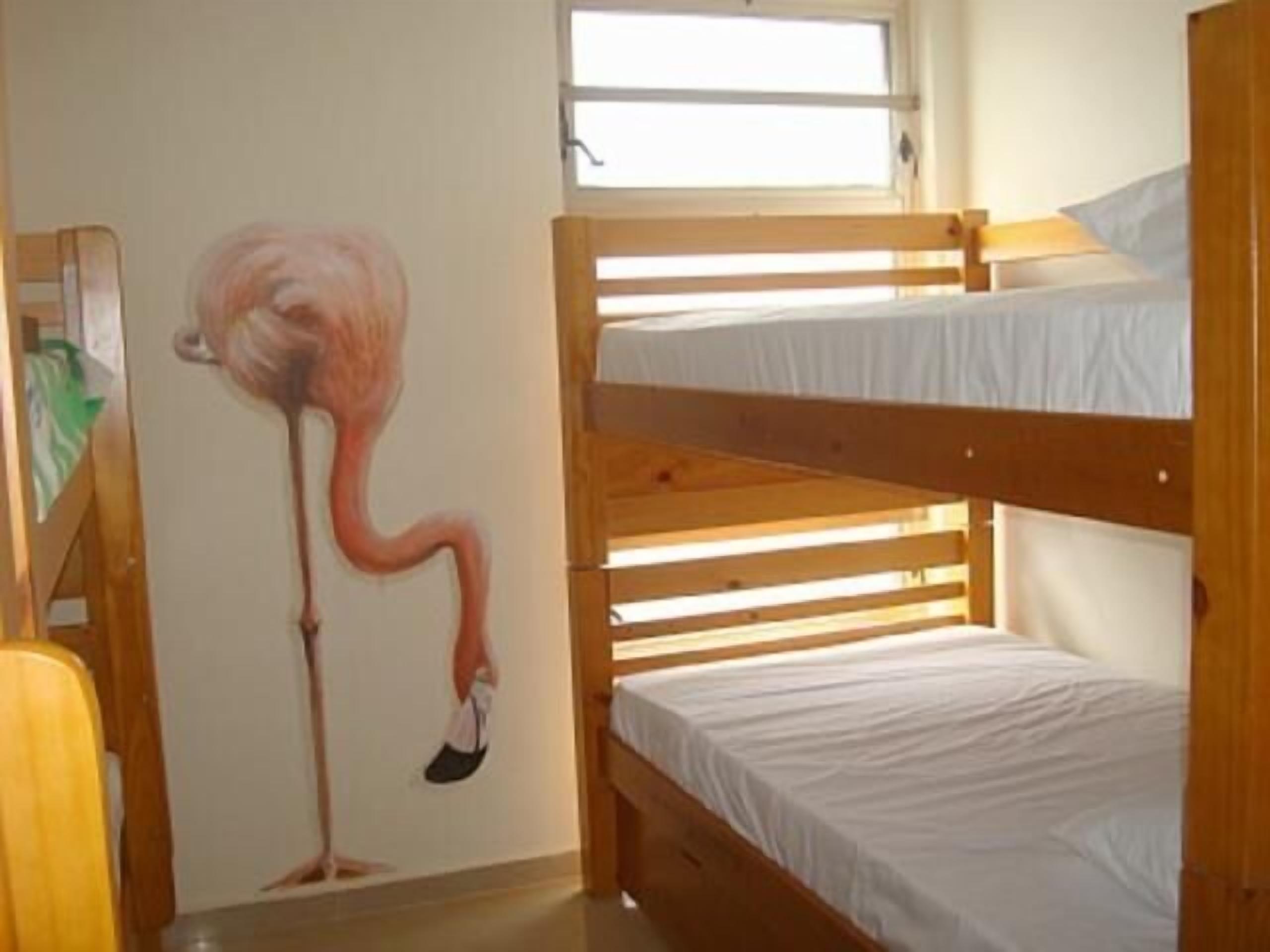 A bunk bed room with a wooden bunk bed and a single bed. There is a window above the bunk bed and a painting of a flamingo on the wall.
