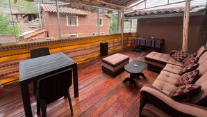 A covered outdoor seating area with a table and chairs, a sofa with cushions, and a view of buildings through large windows. - Casa Intihuatana - Hostel (Pisac)