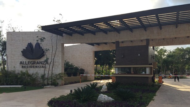 A residential complex entrance with a stone facade, a large flower logo, and a covered walkway. - Residencial Allegranza 628 by Sinbad (Playa del Carmen)