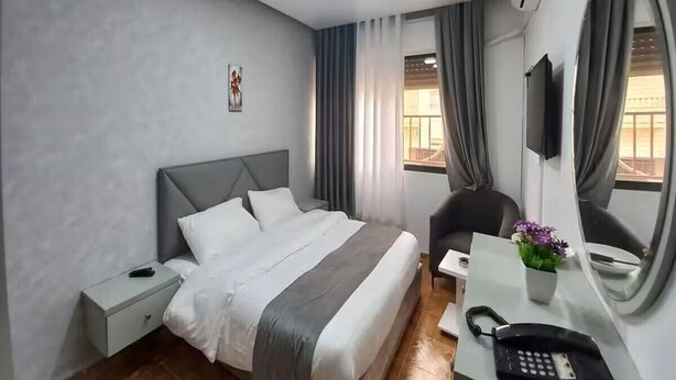 A hotel room with a bed, bedside table, a desk with a phone, a chair, and a TV mounted on the wall. - Al-Nujoom Hotel Suites (Amman)