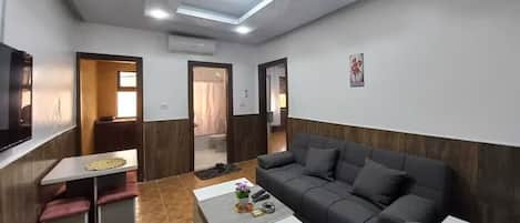 A modern living room with a sofa, a coffee table, and a TV. There is a small table with a vase and a picture frame. The room has wooden paneling and a ceiling with recessed lighting.