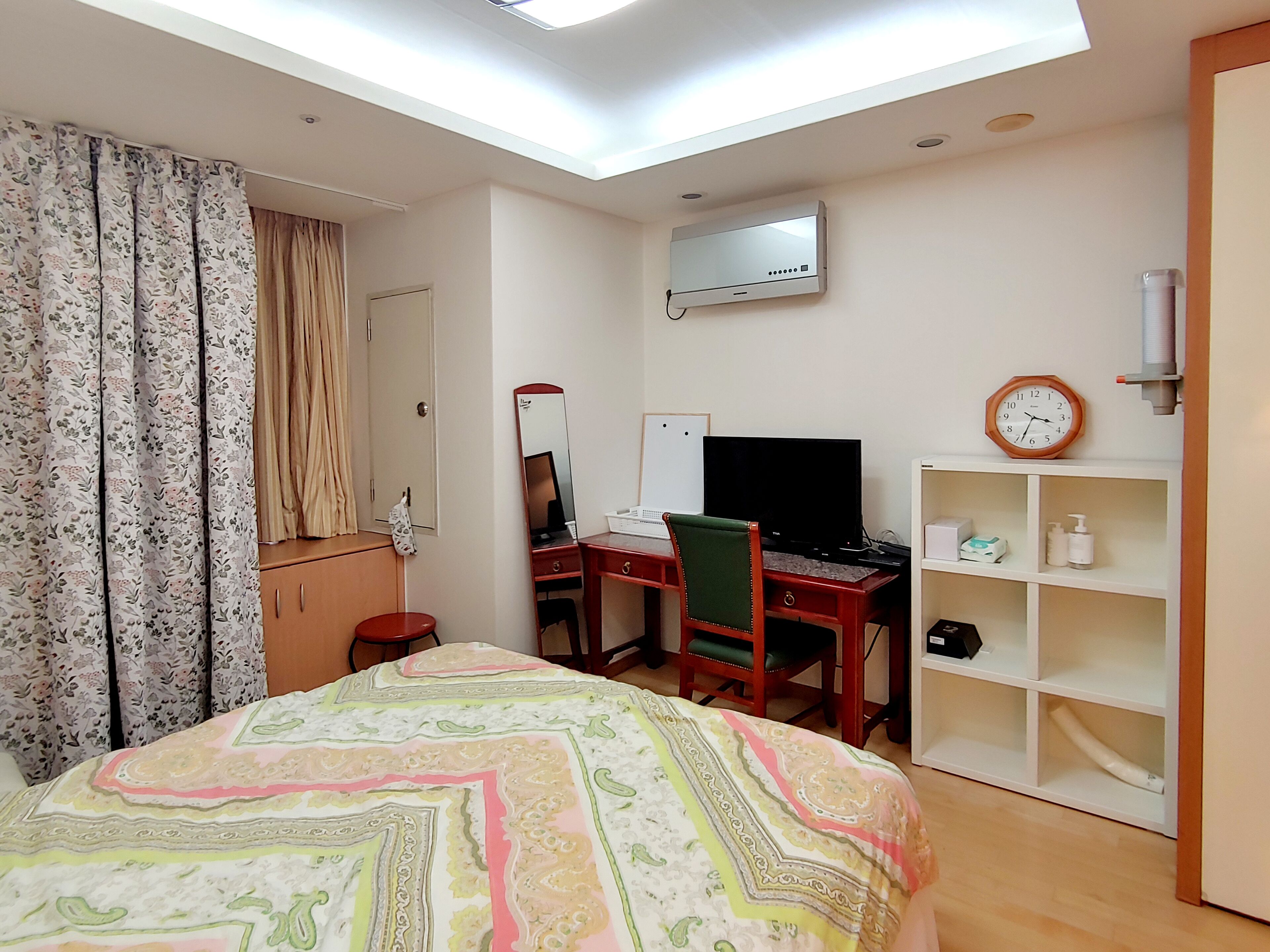 A hotel room with a bed, a desk with a computer, a TV, a mirror, a wardrobe, and a shelf with toiletries.