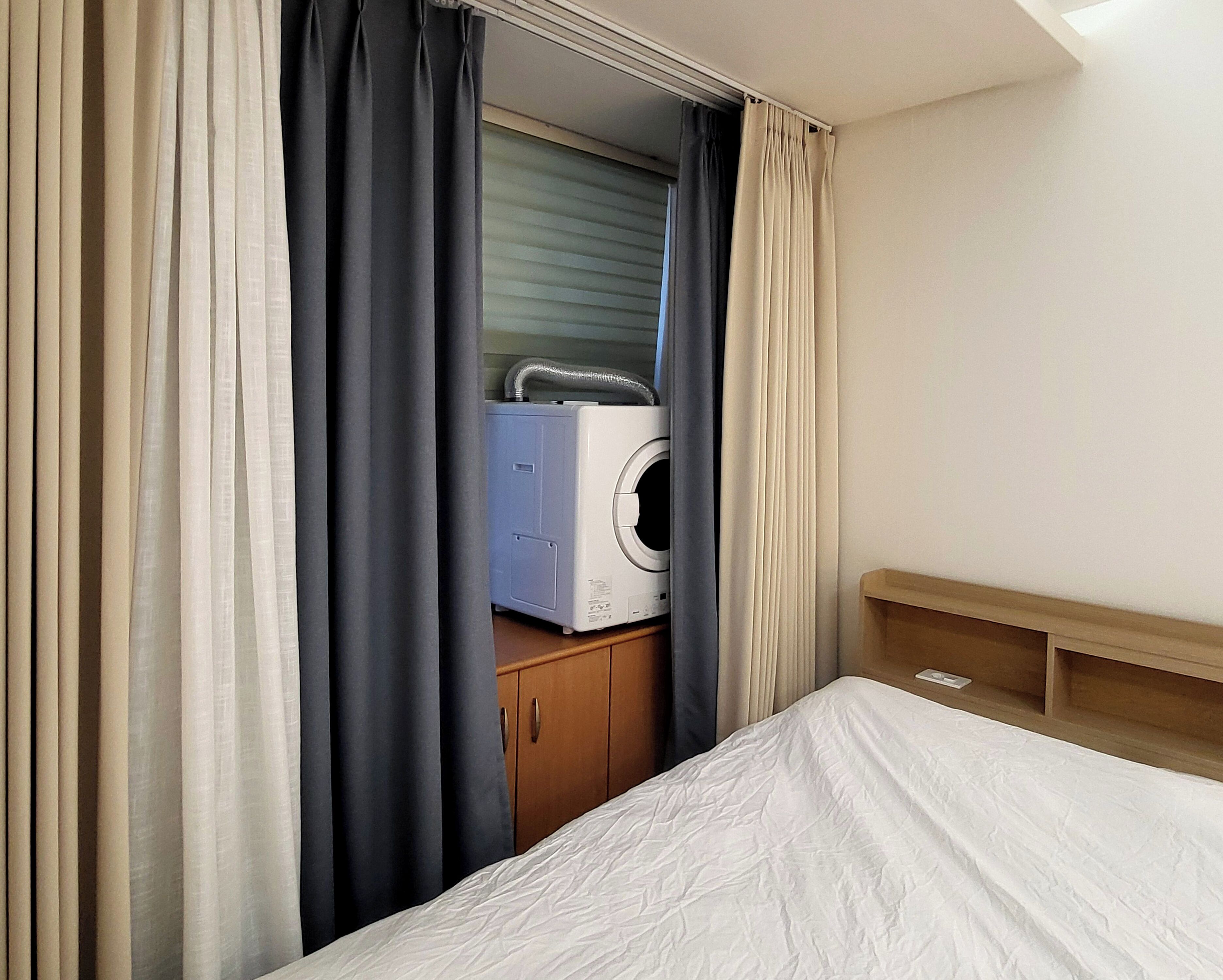 A hotel room with a bed, a wooden headboard, a wardrobe, and a white appliance, possibly a washing machine, in the background.