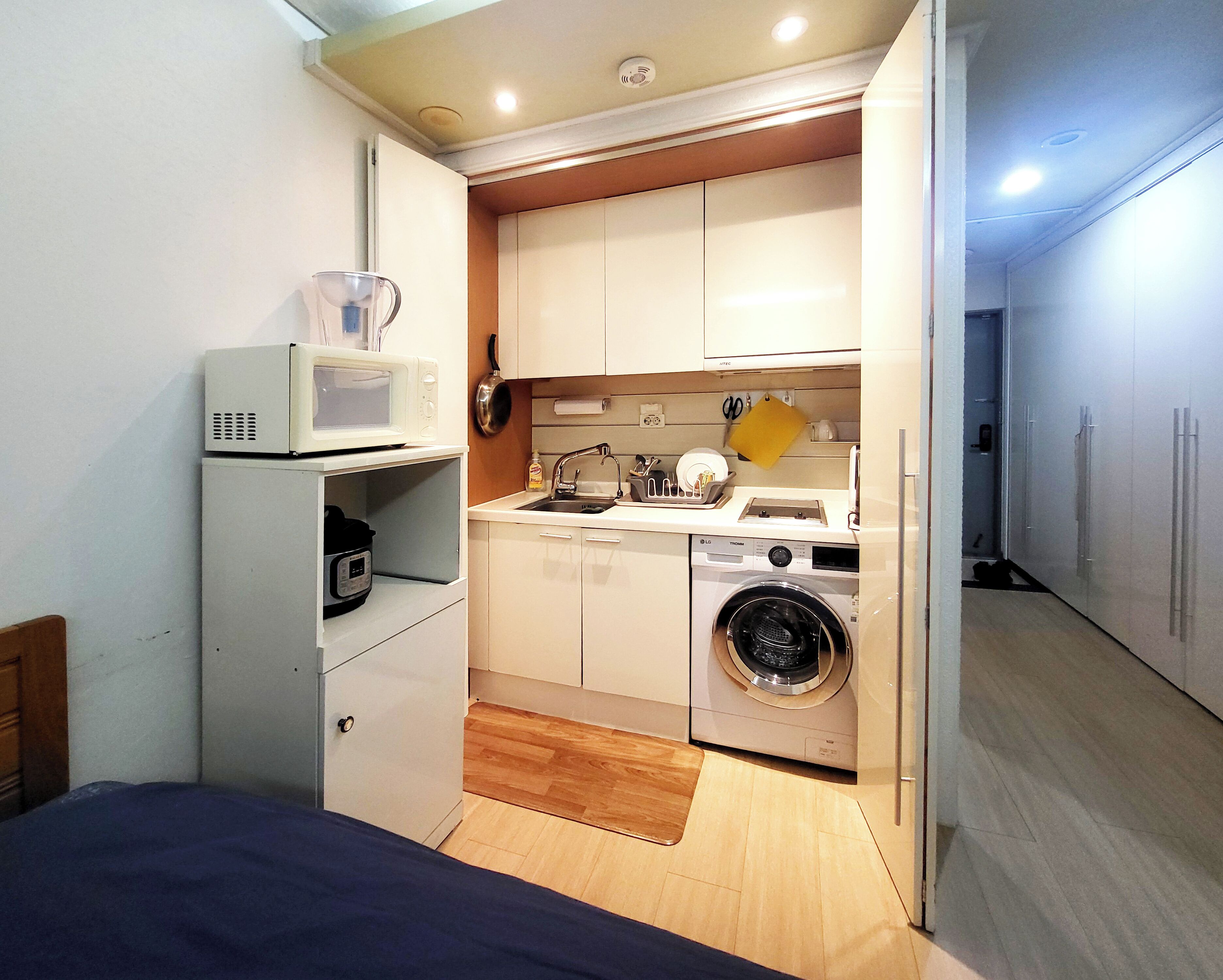 A compact kitchen with a microwave, sink, and washing machine, and a bed in the adjacent room.