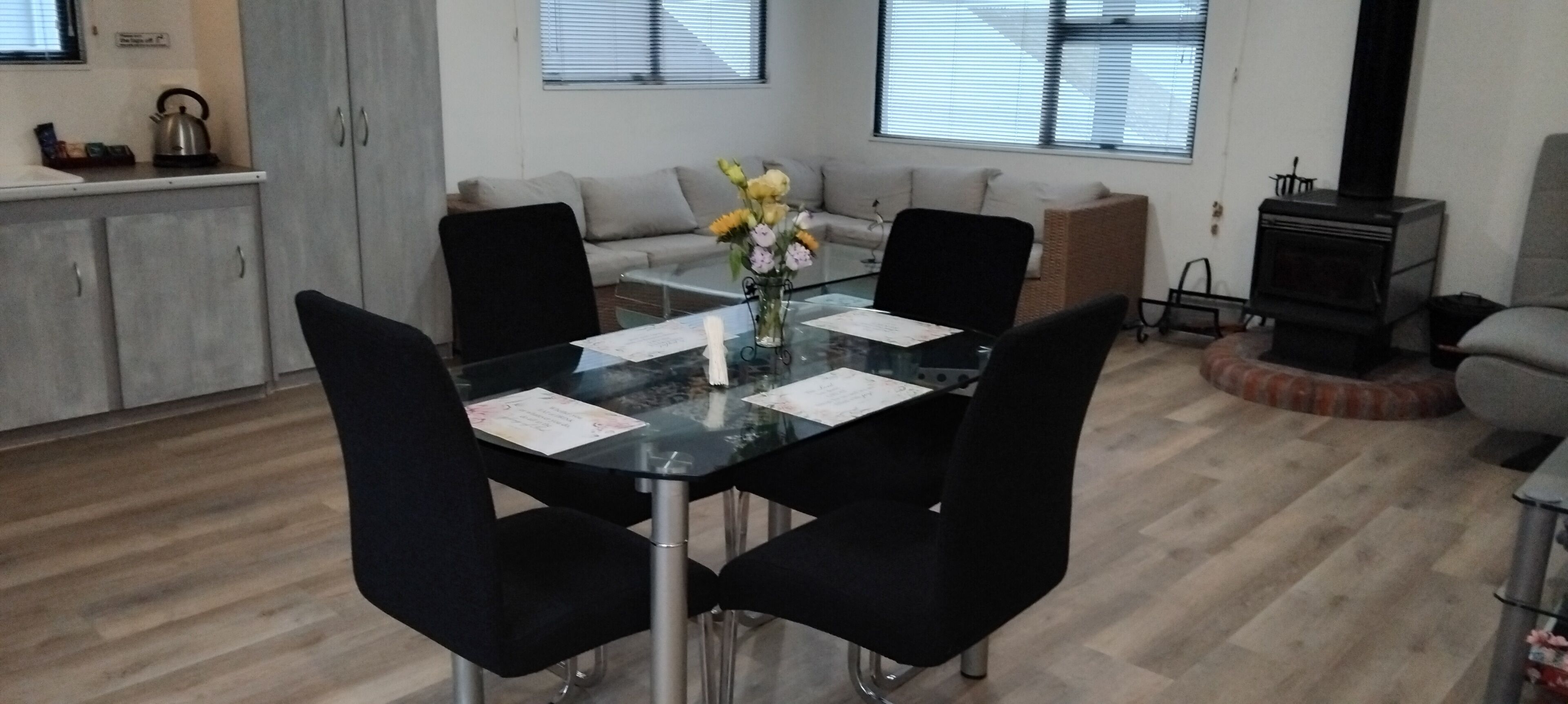 A dining area with a glass table, black chairs, a sofa, a fireplace, and a kitchen area with cabinets and a kettle.
