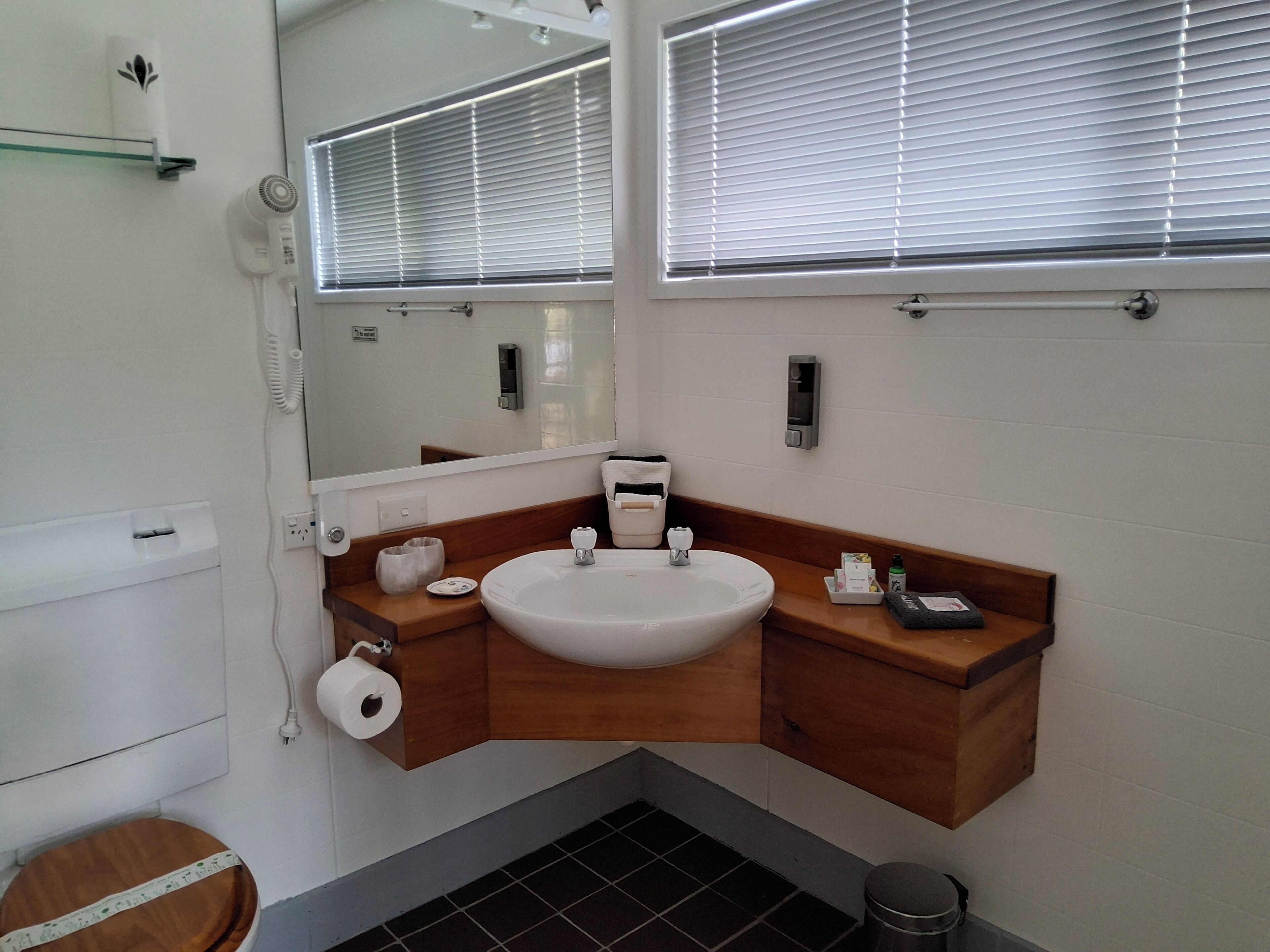 Un baño con lavamanos blanco, tocador de madera, espejo, inodoro y cubo de basura.