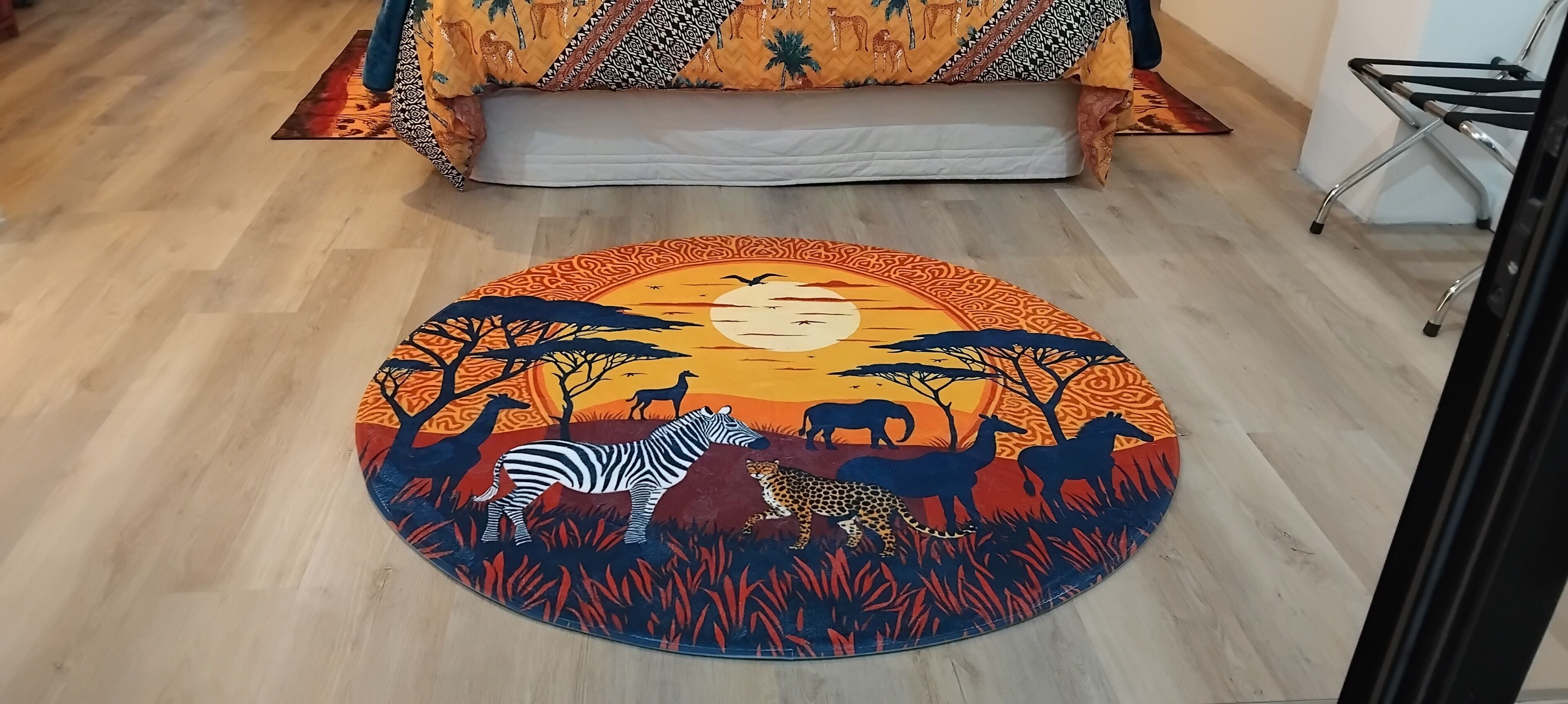 A round rug with a savannah scene, a bed with patterned bedding, and a chair.