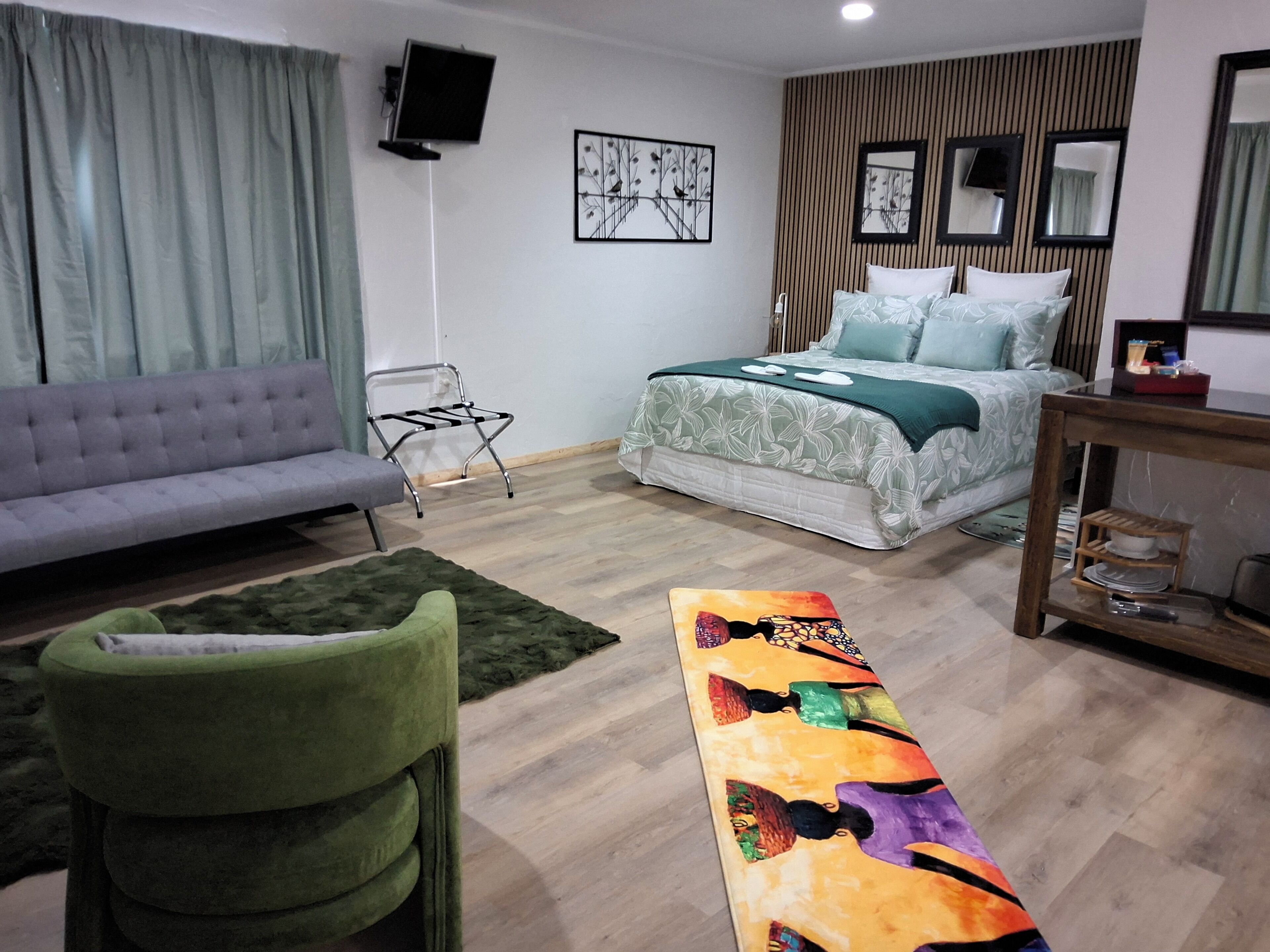 Un dormitorio moderno con sofá, cama con colcha floral, sillón verde y banco colorido.