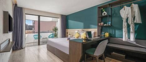 A hotel room with a bed, desk, chair, and a TV. There is a balcony with a view of buildings and a plant.