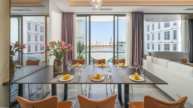 A modern dining area with a large table set for six, large windows offering a city view, and a sofa in the background. - Satya Da Nang Hotel Han Market (Da Nang)